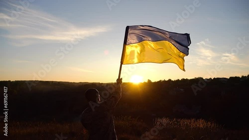 Young man in military uniform waving flag of Ukraine against sunset at background. Male ukrainian army soldier lifted national banner at countryside. Victory against russian aggression. End of war photo
