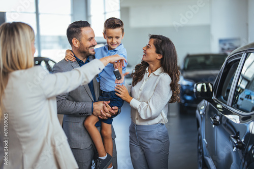 Happy family choosing new car, salesman showing them luxury auto at automobile dealership store. Customers selecting vehicle, consulting manager at modern showroom shop photo