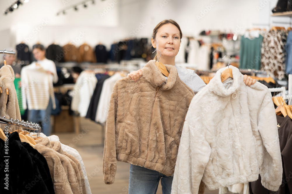 Pleased middle-aged woman buyer choosing short fur coat in clothing shop with large assortment
