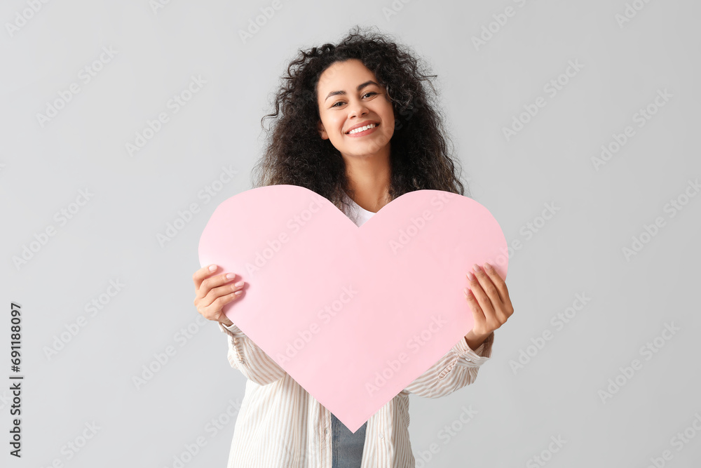 Beautiful young African-American woman with pink paper heart on grey background