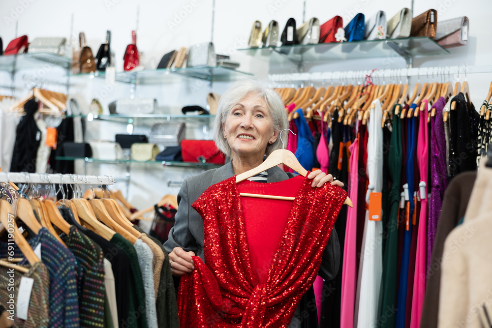 Pleased mature woman choosing evening dress in clothing shop with large assortment