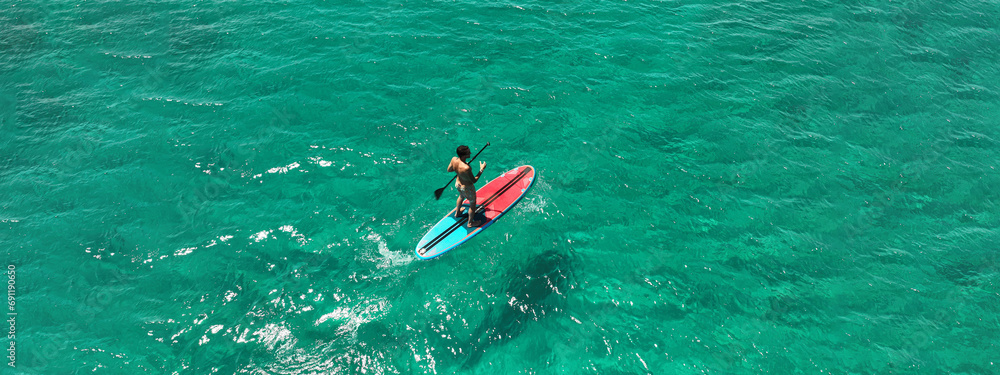 Aerial drone ultra wide panoramic photo of fit man practise SUP or Stand Up Paddle board in tropical exotic bay with emerald calm sea