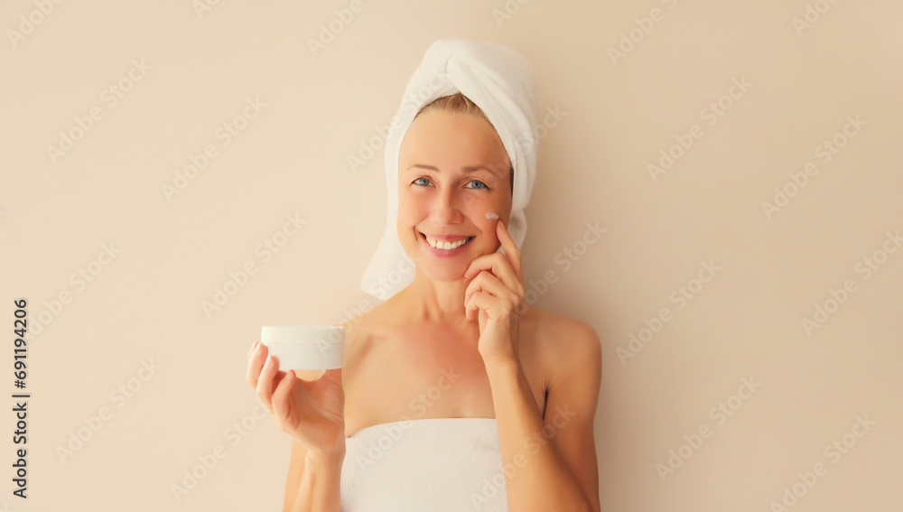 Natural beauty portrait of happy smiling caucasian middle-aged woman touches her clean skin applying face cream while drying wet hair with white wrapped bath towel on her head after shower in morning Natural beauty portrait of happy smiling caucasian middle-aged woman touches her clean skin applying face cream while drying wet hair with white wrapped bath towel on her head after shower in morning