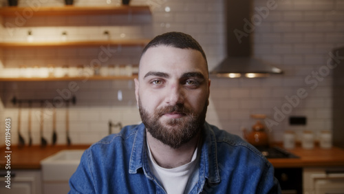 Bearded young adult business man looking at the camera and listening, making conference business call in the kitchen. Video call event by work or study concept	