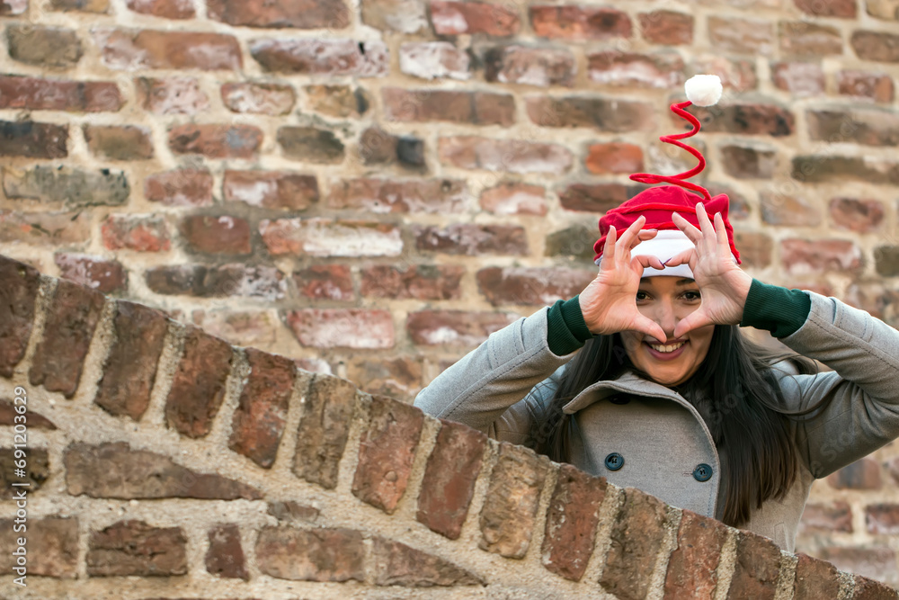 Attractive young brunette in grey coat and santa claus hat near the brick wall making heart shape with her fingers Attractive young brunette in grey coat and santa claus hat near the brick wall making heart shape with her fingers