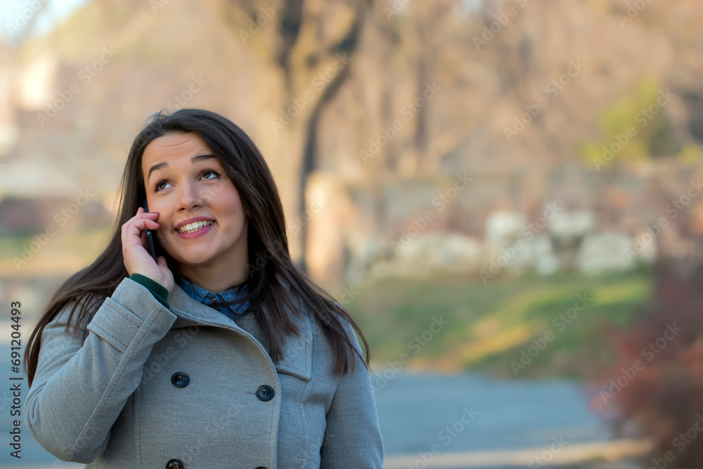 Beautiful young brunette in grey coat walking down the street and talking over phone. City life Beautiful young brunette in grey coat walking down the street and talking over phone. City life