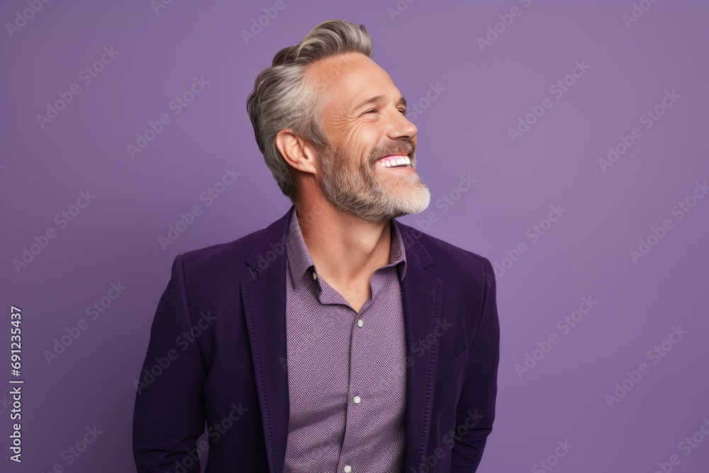Handsome middle-aged man with gray hair and beard is laughing and looking aside while standing against purple background Handsome middle-aged man with gray hair and beard is laughing and looking aside while standing against purple background