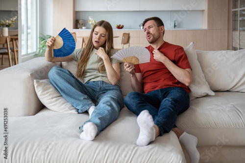 Unhappy couple sitting on sofa waving with hand fans because of broken conditioner, absence of ventilator, air blower. Wife and husband feeling unwell, dehydrated, having difficult breath, headache