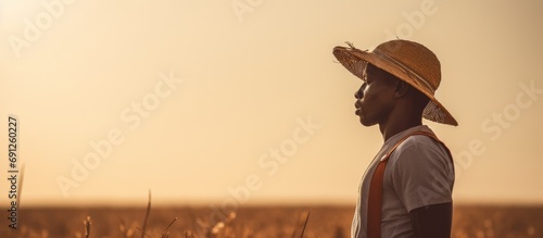 A young African farm worker, carrying a hoe and shielding his eyes from the bright sun, surveys his field.