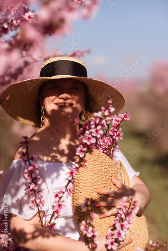 Woman blooming peach orchard. Against the backdrop of a picturesque peach orchard, a woman in a long white dress and hat enjoys a peaceful walk in the park, surrounded by the beauty of nature.