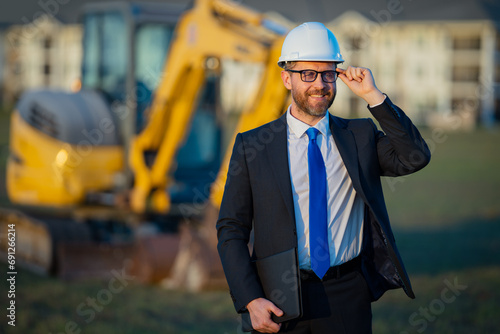 Civil engineer worker at a construction site. Mature engineer worker. Man in suit and hardhat helmet at construction site. Middle aged head civil engineer worker standing outside near excavator.