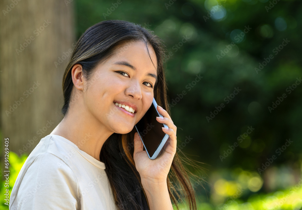Young Asian woman looking at the camera smiling while holding a mobile phone against a blurred background Young Asian woman looking at the camera smiling while holding a mobile phone against a blurred background