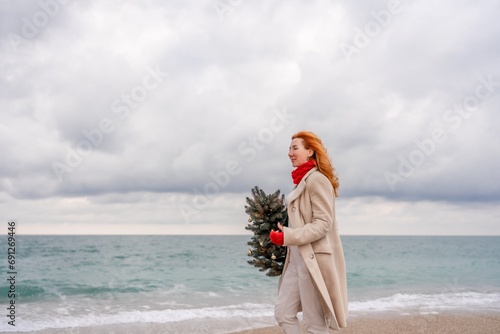 Redhead woman Christmas tree sea. Christmas portrait of a happy redhead woman walking along the beach and holding a Christmas tree in her hands. Dressed in a light coat, white suit and red mittens.
