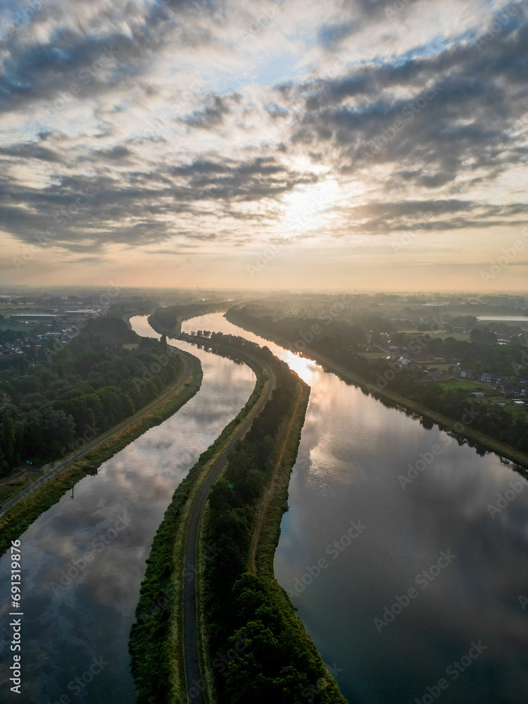 This aerial photograph captures the peaceful essence of a river at sunrise. The water reflects the sky's soft palette of colors while a gentle mist hovers above the surface, blending the boundaries