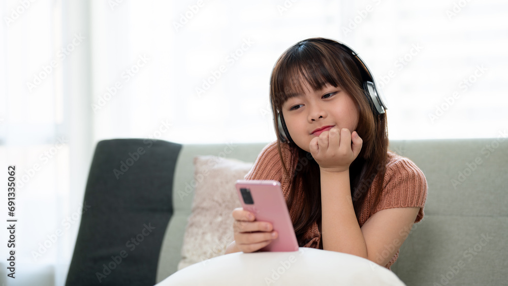 Young adorable Asian girl wearing headphones and using phone, enjoying listening to music on a sofa. Young adorable Asian girl wearing headphones and using phone, enjoying listening to music on a sofa.