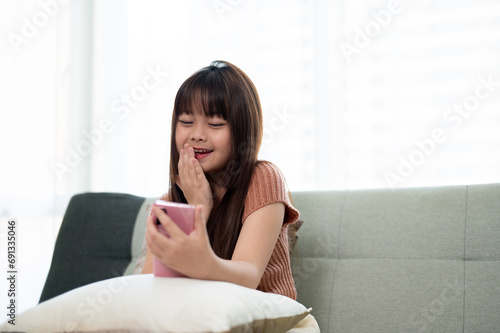 Young cute Asian girl using her smartphone on a sofa in a living room, watching kid cartoons.