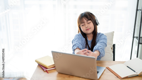 A cute young Asian girl is feeling tired, stretching her arms while studying online at home.