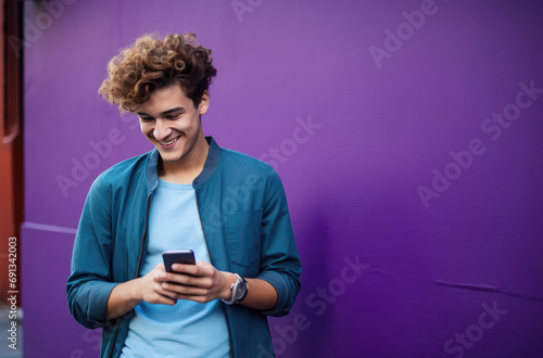 a young man is smiling and holding his smartphone next to a purple wall