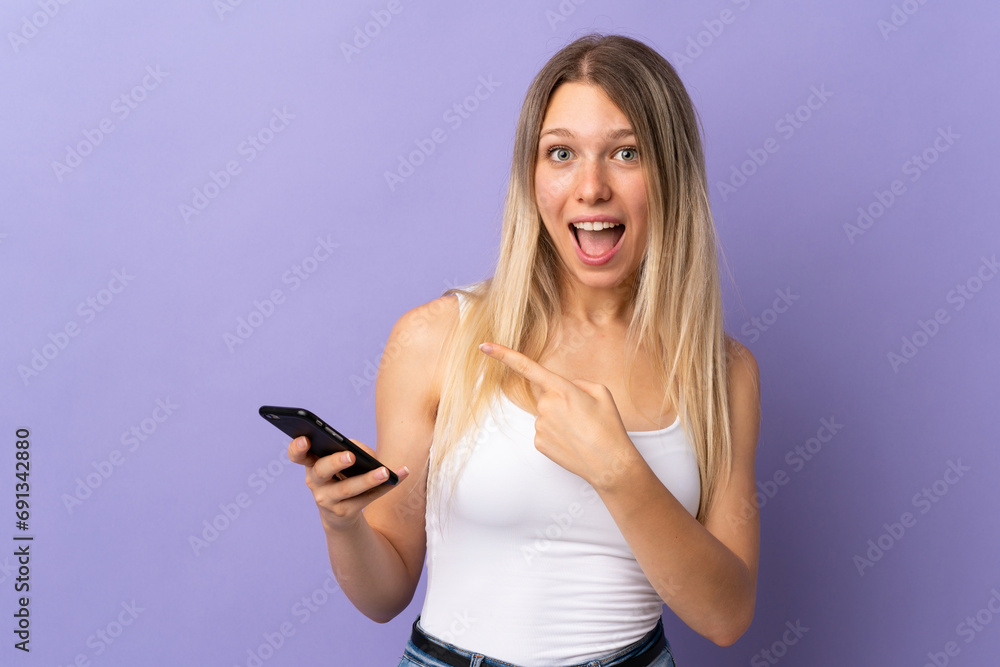 Young blonde woman using mobile phone isolated on purple background pointing finger to the side Young blonde woman using mobile phone isolated on purple background pointing finger to the side