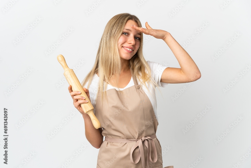 Young woman holding a rolling pin isolated on white background saluting with hand with happy expression Young woman holding a rolling pin isolated on white background saluting with hand with happy expression