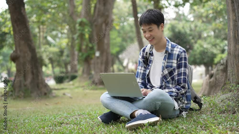 Asian male student reading a book or textbook outdoors under a tree using a laptop. university student Asian male student reading a book or textbook outdoors under a tree using a laptop. university student