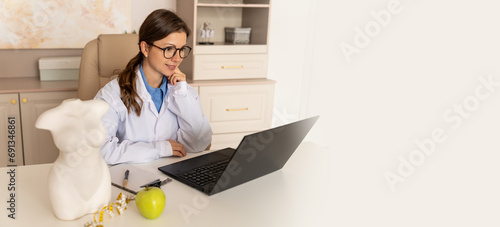 Online consultation with nutritionist banner. Wellness clinic web line. Young beautiful smiling nutritionist working on laptop and writing diet plan for patient and consulting online. Copy space