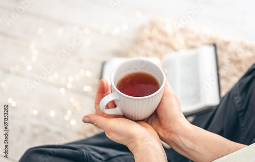 A cup of tea in female hands close-up, book on a carpet on the floor in a room.