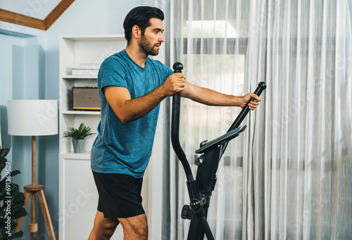 Athletic and sporty man running on elliptical running machine during home body workout exercise session for fit physique and healthy sport lifestyle at home. Gaiety home exercise workout training. photo