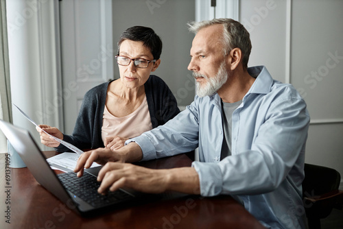 Senior couple talking about family business issues while sitting at table in front of laptop at home photo