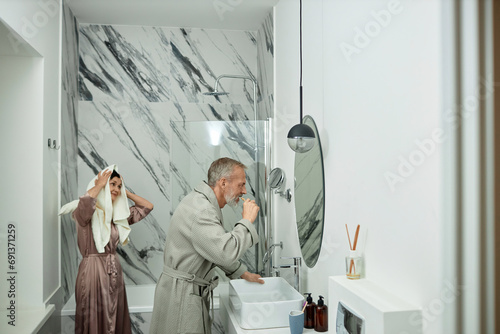 Wife drying hair with towel looking at husband brushing his teeth photo