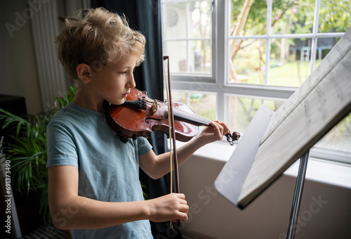 Elementary boy practicing to play violin near window at home photo