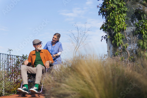 Nurse pushing senior man in wheelchair. Female caregiver and elderly man enjoying a warm autumn day in nursing home. © Halfpoint