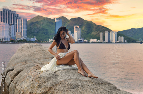 Young ukrainian woman in bikini and white dress sitting on a stone at the Nha Trang beach with peach fuzz shadows of sunset photo
