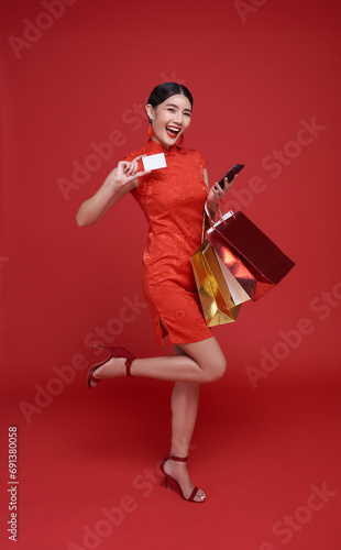 Happy Asian shopper woman wearing traditional cheongsam qipao dress holding credit card and shopping bag isolated on red background. Happy Chinese new year