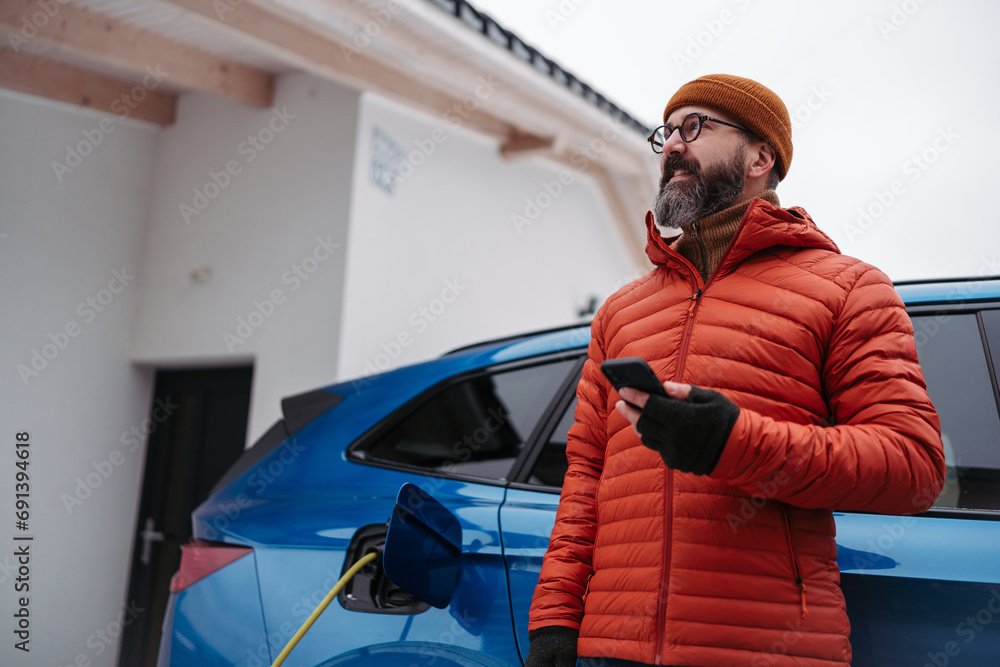 Man charging electric car during cold snowy day, using electric vehicle charging app, checking battery life, energy consumption on smart phone. Charging and driving electric vehicles during winter - obrazy, fototapety, plakaty 