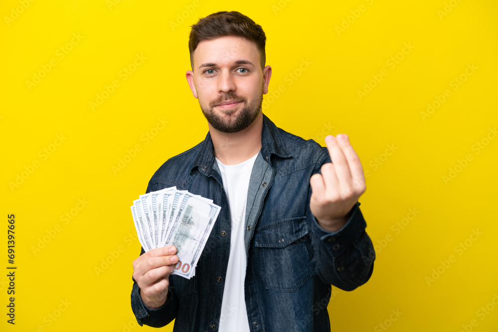 Young caucasian man taking a lot of money isolated on yellow background making money gesture Young caucasian man taking a lot of money isolated on yellow background making money gesture