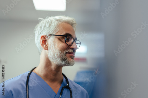 Portrait of confident mature doctor in hospital room. Handsome doctor with gray hair wearing medical scrubs and stethoscope around neck standing in modern private clinic, talking with patient, nurse. photo