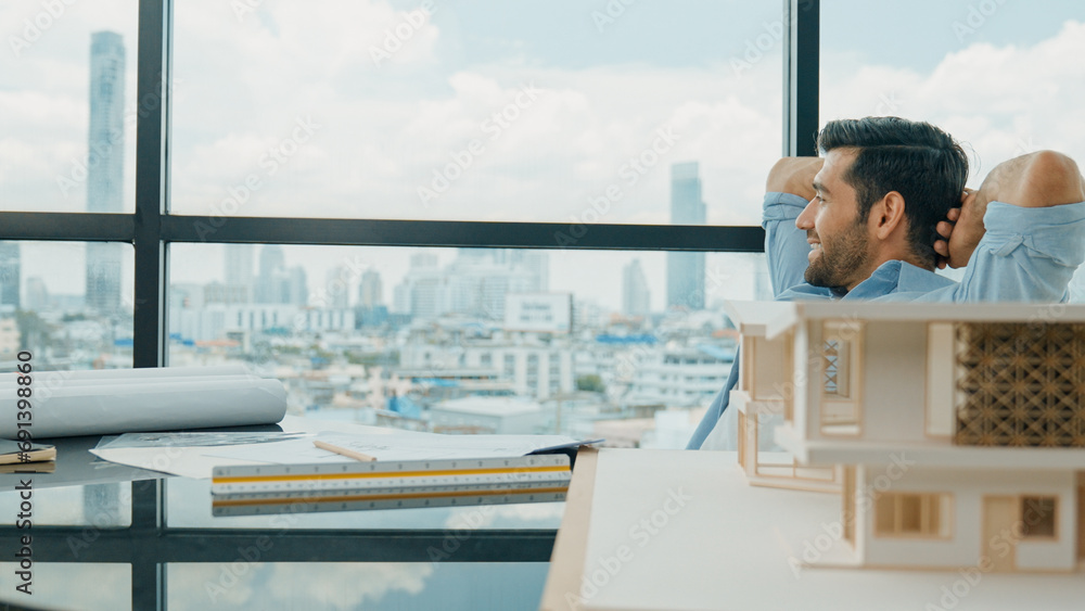 Portrait of relaxed young architect engineer resting and designing while sitting in modern home office. Male worker or manager take a break, hold hands behind head after finish project. Tracery