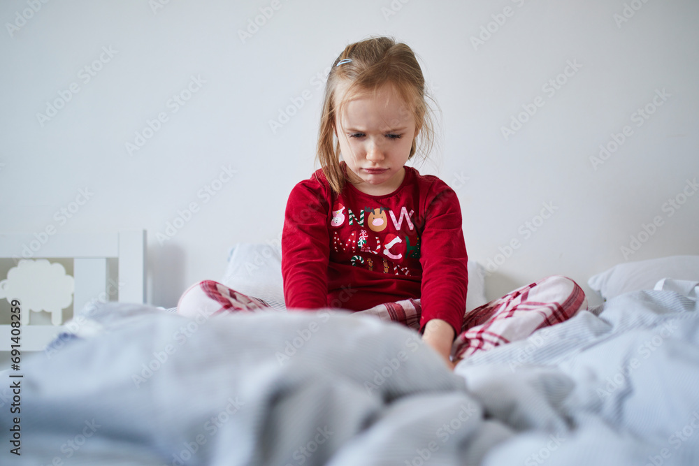 Unhappy preschooler girl wearing Christmas pajamas, sitting on bed Unhappy preschooler girl wearing Christmas pajamas, sitting on bed