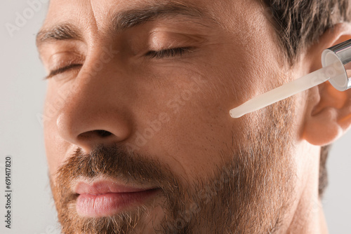 Handsome man applying cosmetic serum onto his face on light grey background  closeup