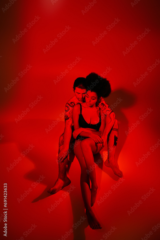 handsome man with tattoos posing with his african american girlfriend in red lights, sexy couple