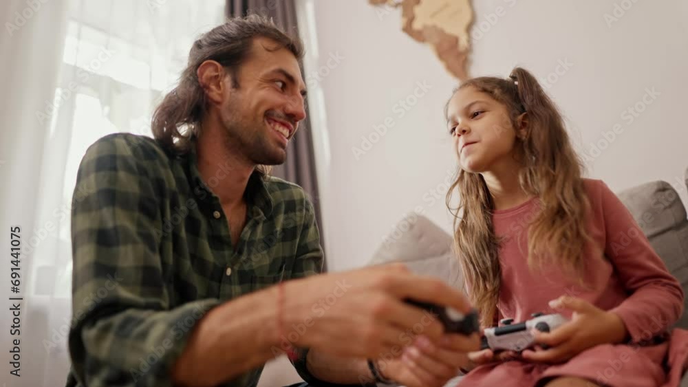 Close-up shot of a brunette man in a green checkered shirt playing and telling how to properly hold a joystick in his hands together with his daughter, a little brunette girl in a pink dress on a gray Close-up shot of a brunette man in a green checkered shirt playing and telling how to properly hold a joystick in his hands together with his daughter, a little brunette girl in a pink dress on a gray