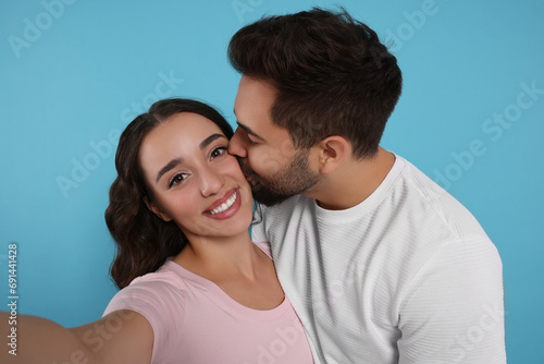 Man kissing his smiling girlfriend while she taking selfie on light blue background © New Africa