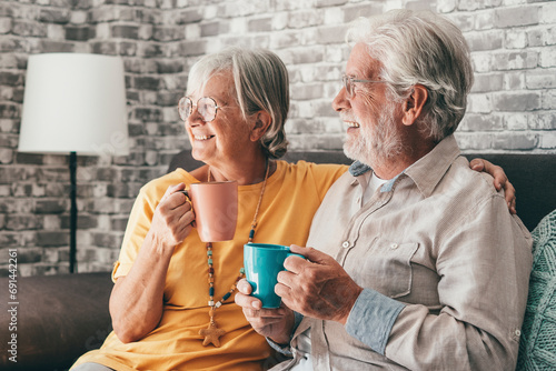 Happy mature 50s husband and wife sit rest on comfortable sofa in living room enjoy tea talking, smiling elderly 60s couple relax on couch at home drink coffee chat speak laugh on leisure weekend. photo