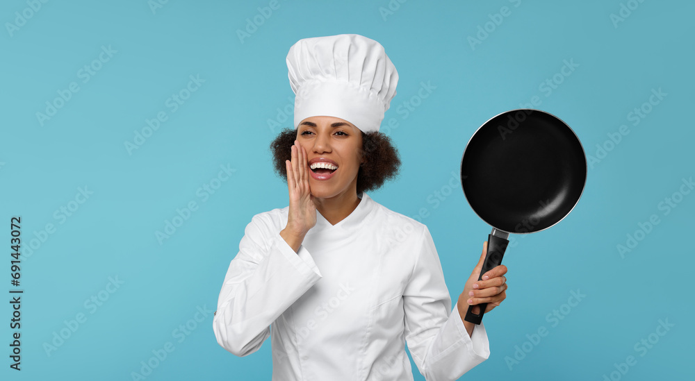 Happy female chef in uniform holding frying pan on light blue background Happy female chef in uniform holding frying pan on light blue background