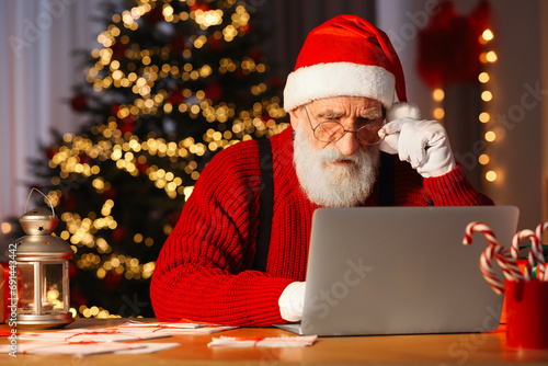 Santa Claus using laptop at his workplace in room with Christmas tree