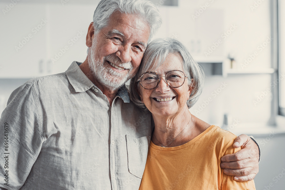 Headshot portrait of smiling elderly 60s husband and wife sit relax on couch hugging cuddling, happy mature old couple rest on sofa in living room embrace look at camera show love and care. Headshot portrait of smiling elderly 60s husband and wife sit relax on couch hugging cuddling, happy mature old couple rest on sofa in living room embrace look at camera show love and care.