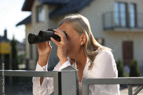 Concept of private life. Curious senior woman with binoculars spying on neighbours over fence outdoors photo