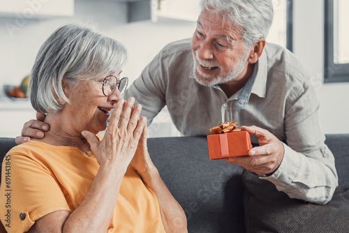 Smiling senior husband making surprise giving gift box to amazed wife, happy aged woman get unexpected present from spouse spending romantic weekend or celebrating anniversary together. photo