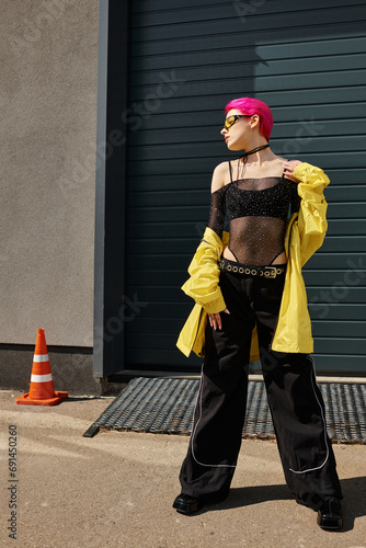 pink haired young woman in sunglasses and trendy outfit posing outdoors, street style fashion